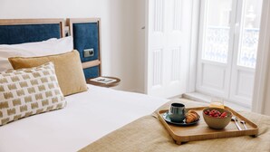 Ein ordentlich bezogenes Bett mit blauem Kopfteil, ein Nachttisch mit einem Buch und ein Frühstücksbrett mit Croissants, Beeren und einer Tasse Tee.
