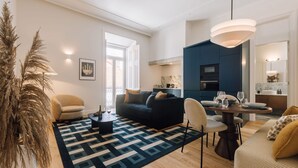 A modern living room with a sofa, chairs, a dining table set for two, and a kitchen in the background. - Ando Living - Abrantes 50 House - 503 (Lisbon)