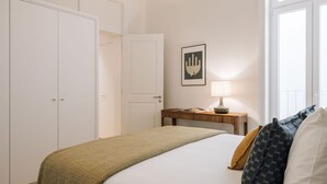 A hotel room with a bed, a bedside table with a lamp, a framed picture on the wall, and a window with curtains. - Ando Living - Abrantes 50 House - 305 (Lisbon)