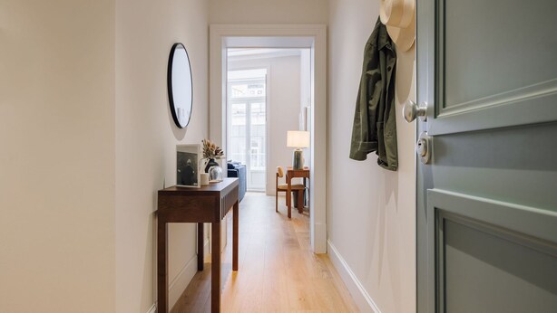 A hallway with a wooden floor, a desk with a lamp, a mirror, and a door with a jacket hanging on it. - Ando Living - Abrantes 50 House - 305 (Lisbon)