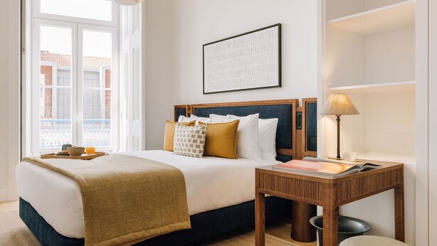 A hotel room with a bed, bedside table, lamp, and a view of the outside. - Ando Living - Abrantes 50 House - 401 (Lisbon)