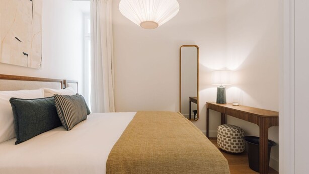 A neatly made bed with pillows, a bedside table with a lamp, a mirror, and a small round ottoman. - Ando Living - Abrantes 50 House - 506 (Lisbon)