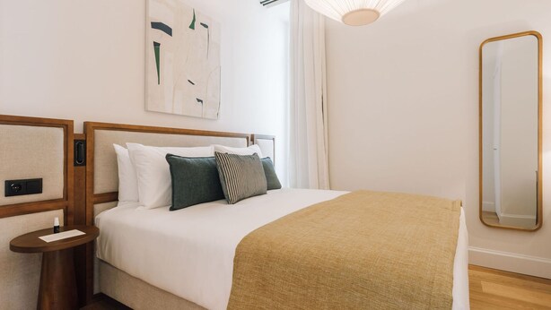 A neatly arranged hotel room with a bed, a bedside table, a mirror, and a painting on the wall. - Ando Living - Abrantes 50 House - 408 (Lisbon)