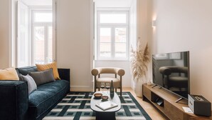 A modern living room with a dark blue sofa, a small round table with a book and glasses, a tall chair, a TV on a wooden stand, and a large window with a view of buildings. - Ando Living - Abrantes 50 House - 408 (Lisbon)
