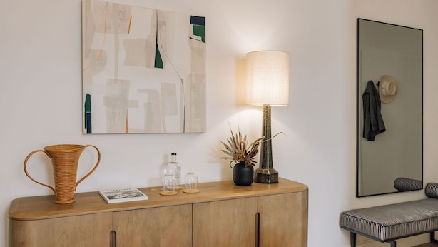 A wooden console table with a woven vase, a lamp, and a small plant. A framed abstract painting and a mirror are on the wall above. - Ando Living - Abrantes 50 House - 402 (Lisbon)