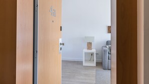 Una habitación con puerta de madera, una mesita de noche blanca, un sofá gris y una lámpara fijada a la pared.