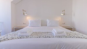 Una cama bien hecha con sábanas blancas y dos almohadas, una manta blanca y dos toallas dobladas sobre un tapete tejido. Hay dos mesitas de noche con lámparas a cada lado de la cama.