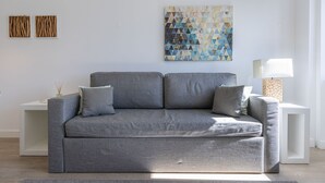A grey sofa with two cushions, a white side table with a lamp, and a geometric artwork on the wall. - Garcia 28 Apartment 1A (Lisbon)