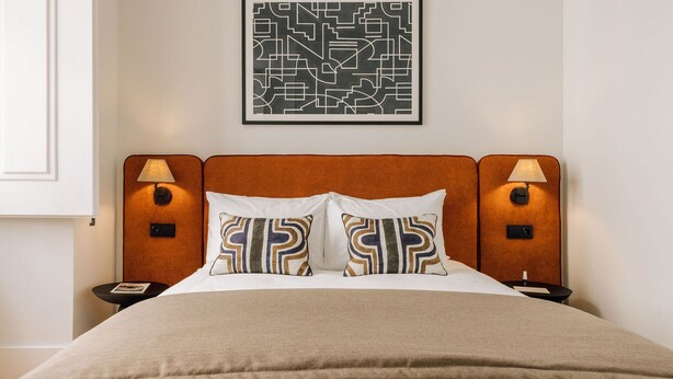 A hotel room with a bed, two bedside lamps, a nightstand, a framed abstract artwork, and a beige throw blanket. - Ando Living - Corpo Santo House - 301 (Lisbon)