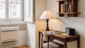 A corner room with a window, a table with a lamp, a chair, a bookshelf, and a radiator. - Ando Living - Corpo Santo House - 301 (Lisbon)