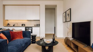 A modern living room with a sofa, a coffee table with wine glasses and a bottle, a TV, and a kitchen in the background. - Ando Living - Corpo Santo House - 301 (Lisbon)