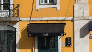 Un edificio con fachada amarilla, toldo negro con letrero que dice "ANDO Living", puerta verde y una pequeña placa que indica "CORPO SANTO HOUSE".