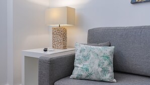 Egy modern nappali, melyben szürke kanapé, fehér dohányasztal, fa tönkből készült lábazattal ellátott lámpa és virágmintás párna található.