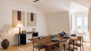 Un coin repas avec une table en bois dressée pour quatre personnes, entourée de chaises en osier. Une lampe se trouve sur une petite table d’appoint noire, et des œuvres d’art ornent le mur.