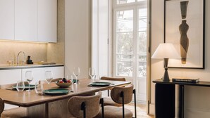 Un espace repas moderne avec une table en bois dressée pour quatre personnes, un saladier de fruits et un livre intitulé « Maisons pour notre époque 2 » posé sur une table d’appoint.