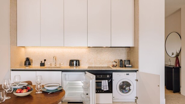 A modern kitchen with white cabinets, a built-in oven, a washing machine, and a dining area with a table set for two. - Ando Living - Corpo Santo House - 501 (Lisbon)