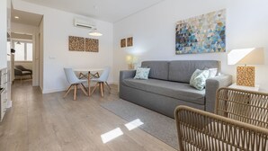 Egy modern nappali, melyben egy szürke kanapé, két világoskék szék, egy étkezőasztal és egy fonott szék található. A falakon képek vannak, és a mennyezetről világít egy lámpa.