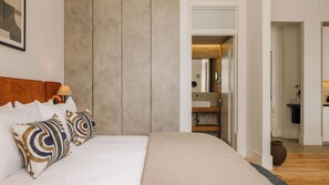 Un dormitorio con cama, almohadas, una mesita de noche y una lámpara. Hay una puerta que da a un baño con espejo y lavamanos.