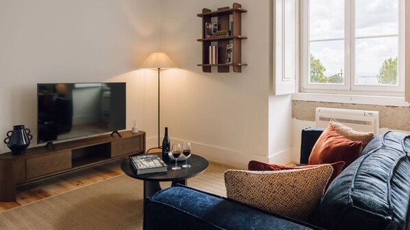 Una sala de estar con televisor, una lámpara, una mesita con copas de vino, una estantería y una ventana con vistas.