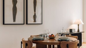 A dining area with a round table set for four, surrounded by wicker chairs. There are two framed abstract paintings on the wall above the table. - Ando Living - Corpo Santo House - 101 (Lisbon)