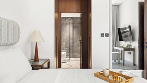 Une chambre d’hôtel avec un lit, une table de chevet avec une lampe, un plateau avec le petit-déjeuner, une télévision et une salle de bain visible à travers une porte en verre.
