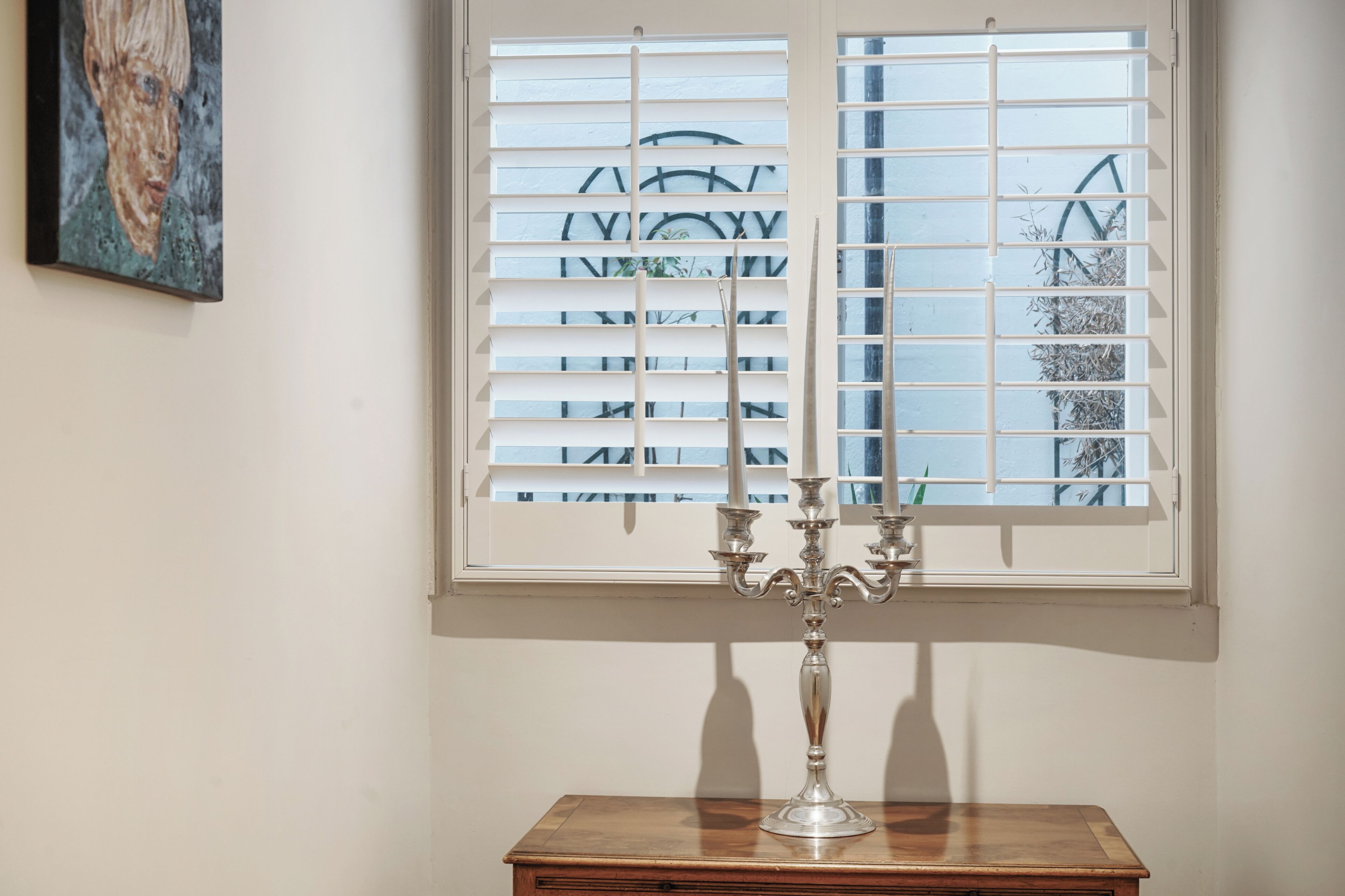 Una ventana con persianas blancas, un candelabro sobre una mesa de madera y un cuadro en la pared.