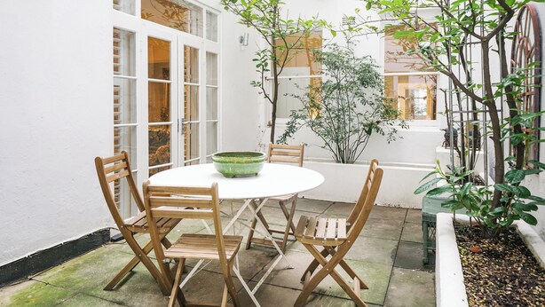 A round wooden table with chairs, a green bowl, and a potted plant in a white-walled outdoor area with a tiled floor. - Queen's Gate Gardens Flat (London)
