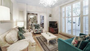 A living room with a sofa, armchair, coffee table, and a television. There is a large window with blinds, a chandelier, and a mirror on the wall. - Queen's Gate Gardens Flat (London)