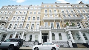 A row of townhouses with a central entrance, a black car parked in front, and a clear blue sky. - Queen's Gate Gardens Flat (London)