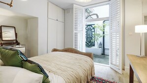 A bedroom with a bed, bedside table, lamp, and a view of the outside. - Queen's Gate Gardens Flat (London)
