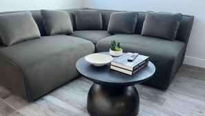 En moderne stue med en grå hjørnesofa, et rundt sort sofabord, en lille potteplante, en hvid skål formet som en snegl, og en bog med titlen "Living Redefined".
