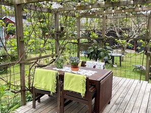 Une terrasse extérieure en bois, avec une table et des chaises, entourée d’une clôture en fil de fer et de verdure.
