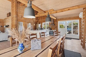 A log cabin interior with a wooden dining table, chairs, hanging pendant lights, a kitchen area, and a large window. - Lovely log house on Møn (Stege)