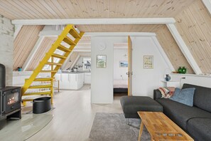 A loft-style room with a wooden ceiling, a staircase, a kitchen area, and a living space with a sofa and a table.