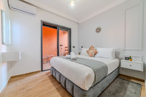 Una camera da letto moderna con un letto grande, un comodino, un tavolino da notte, un condizionatore d'aria a parete e una porta scorrevole in vetro che dà su uno spazio esterno.