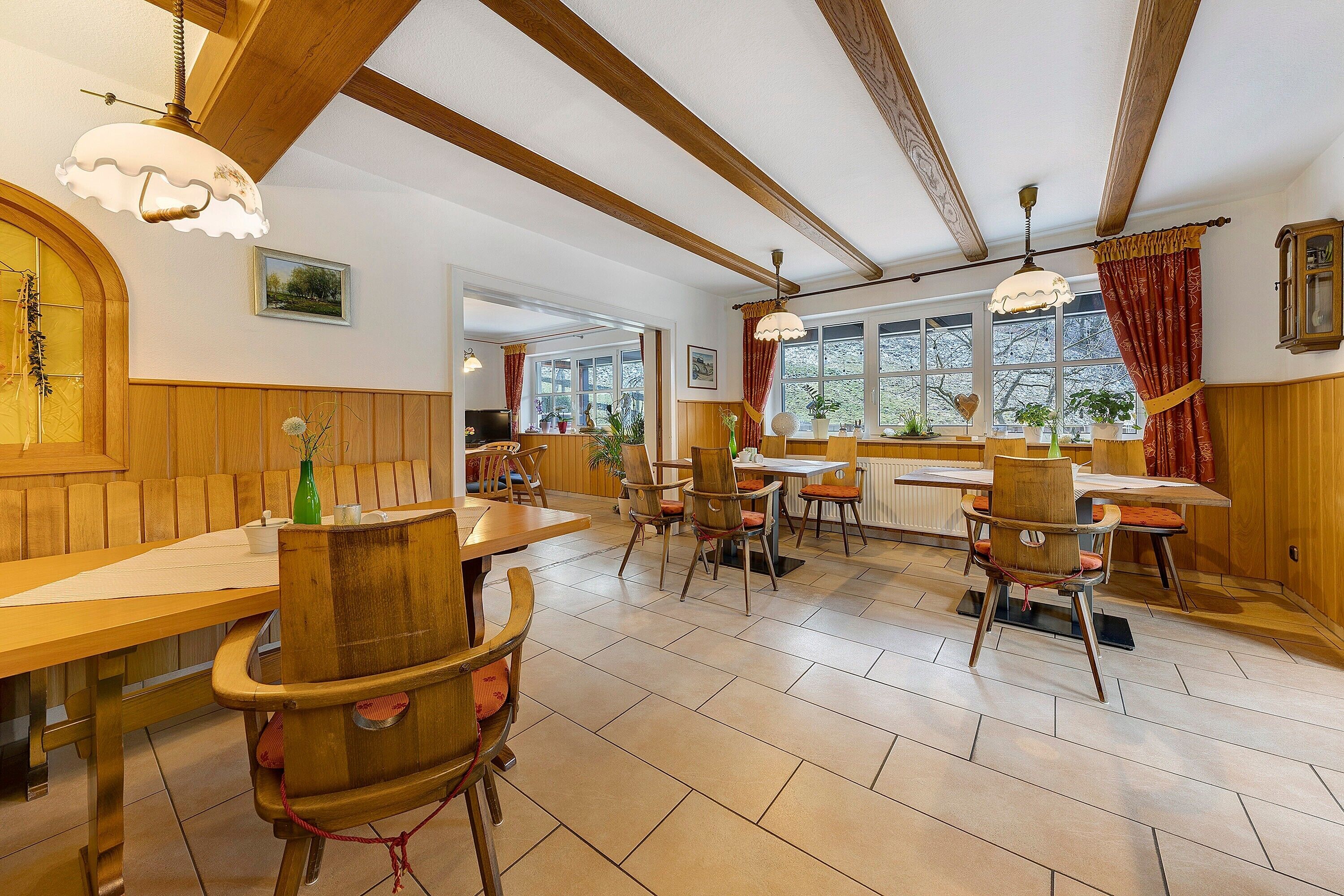 Een eetruimte met houten tafels en stoelen, een kroonluchter en grote ramen.