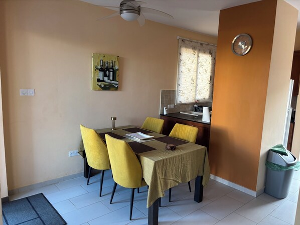 Comedor con una mesa, sillas, ventilador de techo, reloj y ventana con cortinas estampadas.