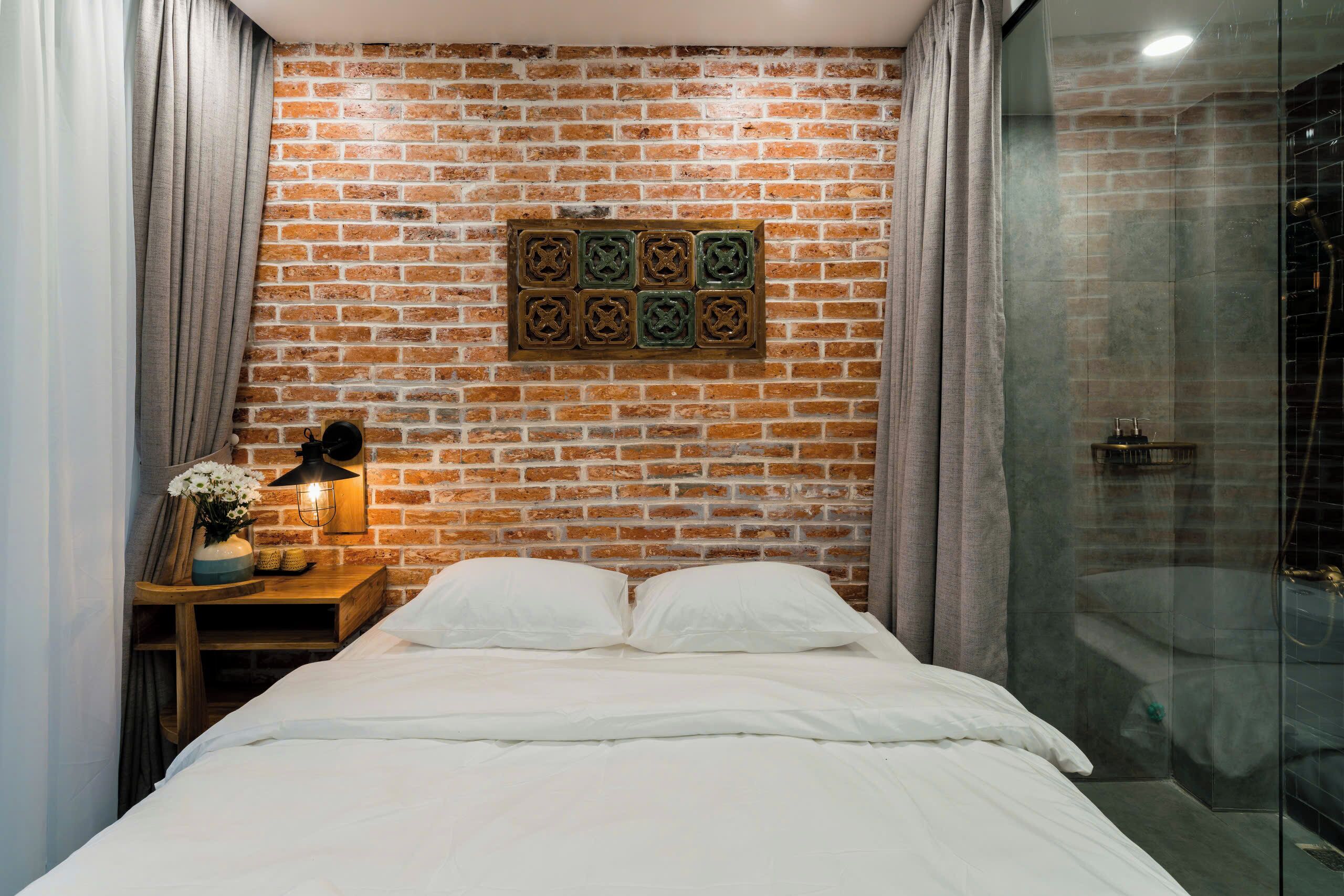 A bedroom with a brick wall, a bed with white bedding, a nightstand with a lamp, and a shower area.