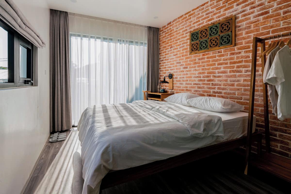 A hotel room with a brick wall, a bed with white linens, a wooden nightstand, a hanging coat rack, and a window with curtains.