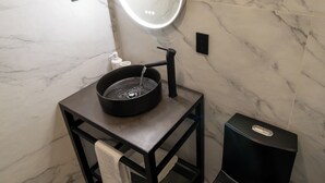 Een badkamer met een marmeren wand, een zwart wastafelmeubel, een ronde spiegel met verlichting, een zwart toilet en een zwarte prullenbak.