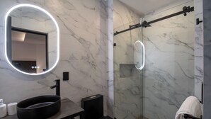 Un baño moderno con pared de mármol, un espejo redondo con iluminación, un lavabo negro y una ducha con mampara de vidrio.