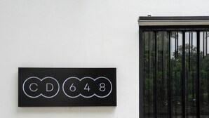 Une plaque noire avec des lettres et des chiffres blancs, fixée sur un mur blanc, à côté d’un portail en métal noir.
