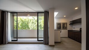 Un vaste espace de vie avec une porte-fenêtre coulissante donnant sur un balcon, une cuisine dotée de meubles de couleur sombre et un téléviseur à écran plat fixé au-dessus du comptoir.