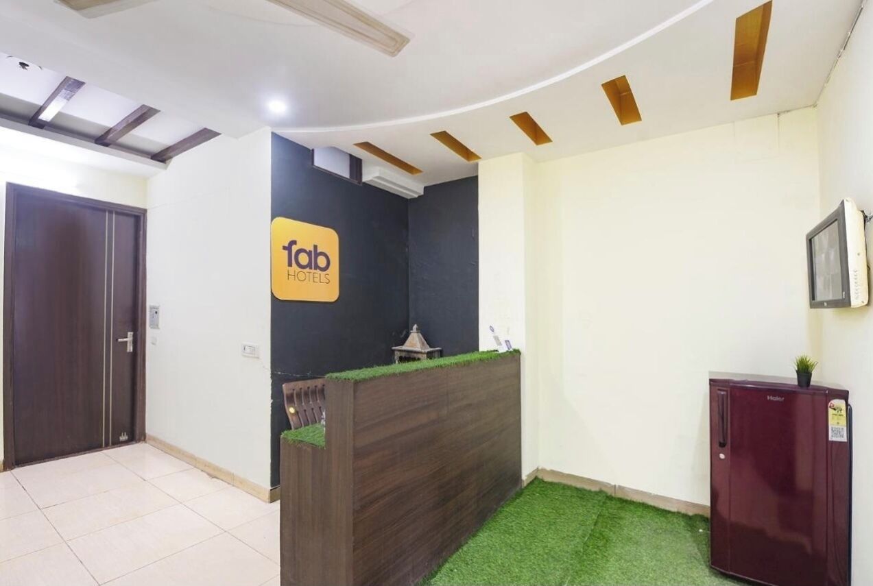 Zona de recepción de hotel con un mostrador oscuro, un cartel que dice 'Tab Hotels', una planta pequeña sobre el mostrador, un refrigerador y una puerta a la izquierda.