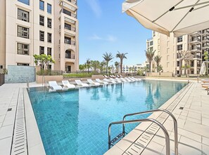 Piscina com espreguiçadeiras e guarda-sol, cercada por um edifício com várias andares e varandas.