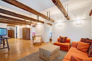 Een moderne woonkamer met zichtbare houten balken, een bank, een fauteuil, een televisie en een gestreept kastje.