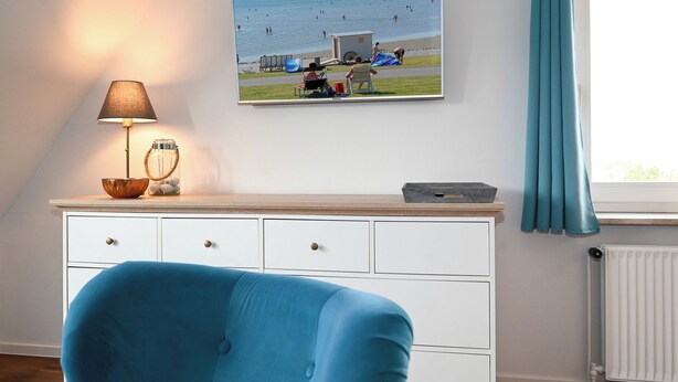 A room with a blue chair, a white dresser, a lamp, and a beach scene painting. - Vacation home Cumulus - Apartment 4 (Büsum)