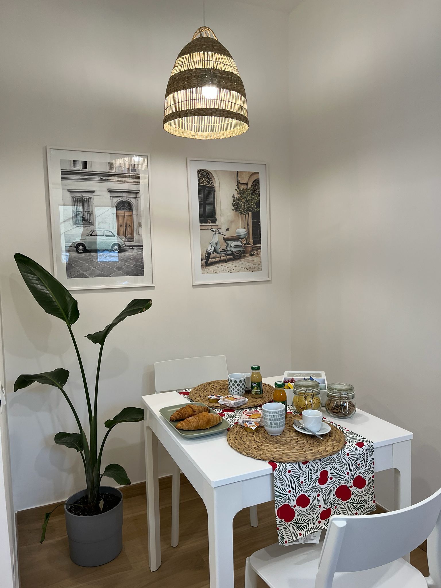 A dining area with a table set for breakfast, including croissants, coffee, and juice. There are two framed pictures on the wall, a potted plant, and a hanging light fixture.