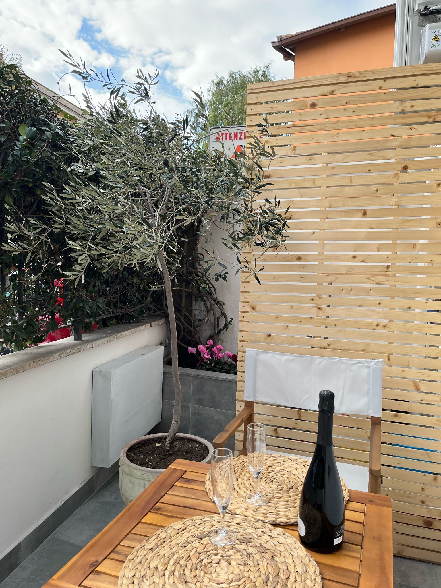 A wooden table with woven placemats, a bottle, and two glasses on a balcony with a potted plant and a wooden fence.