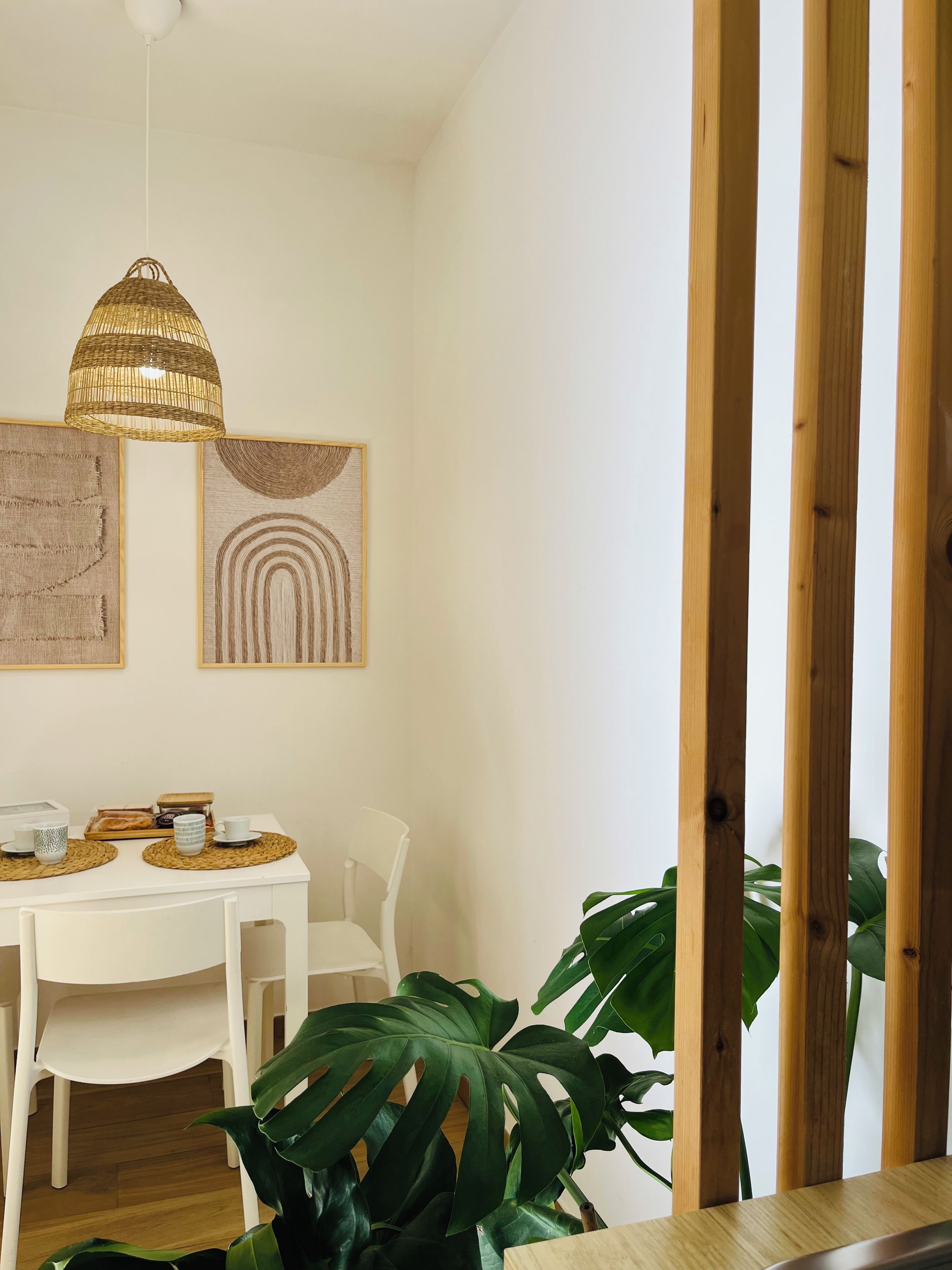 Un coin repas avec une table blanche, deux chaises, un luminaire suspendu en osier et deux œuvres d’art encadrées accrochées au mur.